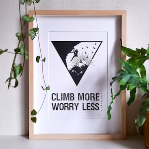 Op de afbeelding: Zwart-witte ingelijste print met een motiverende quote: "Climb More Worry Less". De print toont een silhouet van een persoon die een berg beklimt met een zon en vogels in de lucht.
