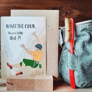 May include: A greeting card with a cartoon illustration of a person rock climbing. The card reads "What the crux, you're how old?!".