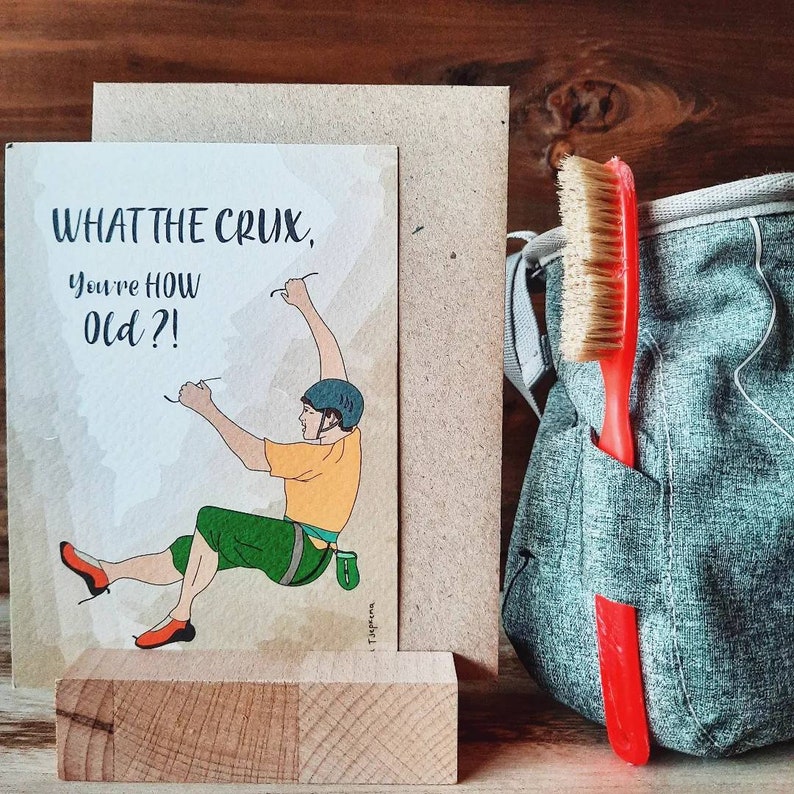 May include: A greeting card with a cartoon illustration of a person rock climbing. The card reads "What the crux, you're how old?!".