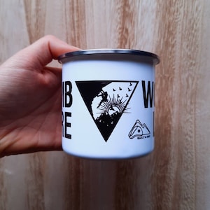 Peut inclure: Mug de camping en émail blanc avec un motif triangulaire noir représentant un grimpeur en silhouette contre un lever de soleil. Le texte "Climb Higher" est sur le côté gauche du mug et "World Is Yours" est sur le côté droit. Le mug a un logo noir qui dit "Rocks & Vibes".