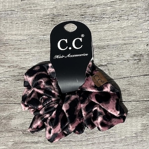 May include: A scrunchie with a pink and black leopard print pattern. The scrunchie is attached to a black tag with the brand name "C.C Hair Accessories" in white lettering.