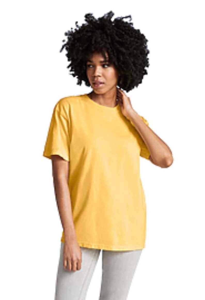 May include: A person wearing a yellow short-sleeved t-shirt and light gray pants. The t-shirt is a solid color and appears to be made of a soft material. The person has dark, curly hair.