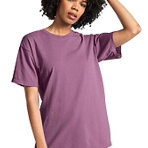 May include: A short-sleeved, mauve-colored t-shirt. The shirt has a crew neck and a relaxed fit. The model is wearing the shirt with white pants. The model has dark, curly hair.