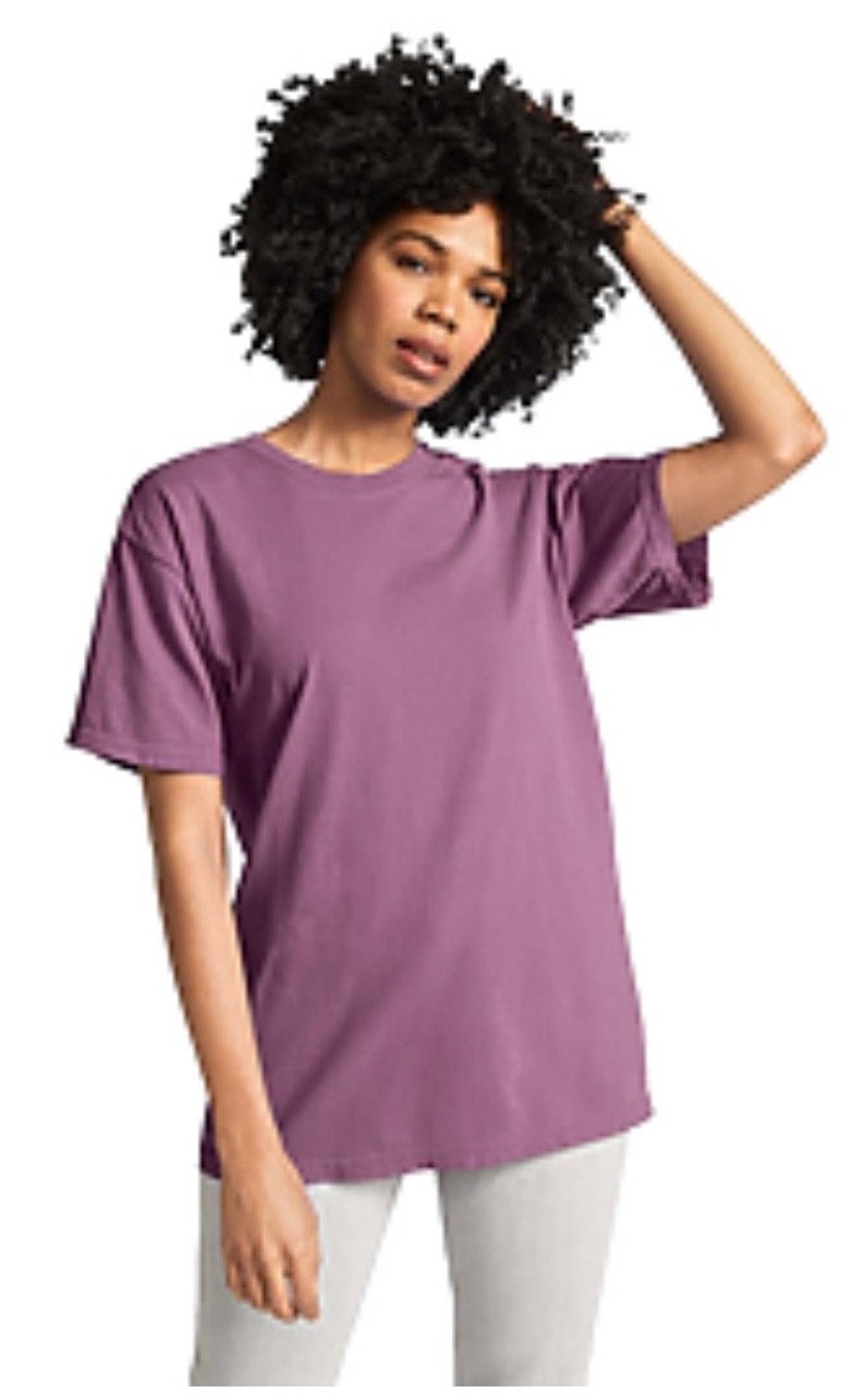 May include: A short-sleeved, mauve-colored t-shirt. The shirt has a crew neck and a relaxed fit. The model is wearing the shirt with white pants. The model has dark, curly hair.