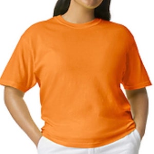 May include: An orange short-sleeved t-shirt is worn with white pants. The t-shirt is a solid color and appears to be made of a soft material. The person is smiling and has their hands in their pockets.