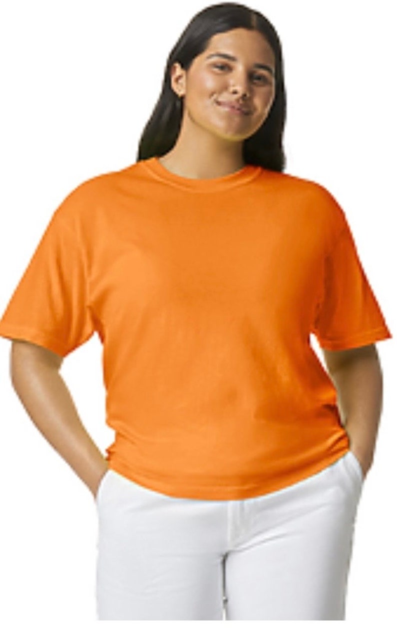 May include: An orange short-sleeved t-shirt is worn with white pants. The t-shirt is a solid color and appears to be made of a soft material. The person is smiling and has their hands in their pockets.