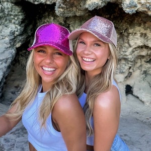May include: Two women wearing sparkly trucker hats, one pink and one rose gold, are smiling at the camera. They are both wearing white tank tops and blue denim shorts.