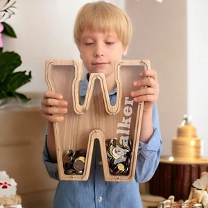 May include: A wooden letter "W" shaped piggy bank with clear acrylic panels, filled with coins. The name "Walker" is printed vertically on the right side. The bank is held by a child wearing a blue shirt.