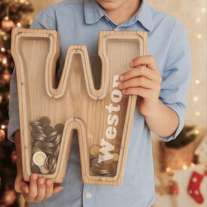 May include: A wooden "W" shaped money box with clear panels, containing coins. The name "Weston" is printed vertically on the side. The money box is held by a person in a blue shirt, with a Christmas tree and star in the background.