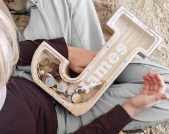 Könnte beinhalten: Ein Sparschwein aus Holz in Form des Buchstabens J mit einem Schlitz für Münzen. Das Sparschwein ist mit dem Namen "James" personalisiert.