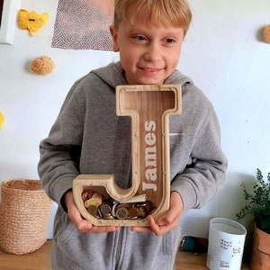 Op de afbeelding: Een houten spaarpot in de vorm van de letter J met de naam "James" erop gegraveerd. De spaarpot zit vol met munten en wordt vastgehouden door een kind.