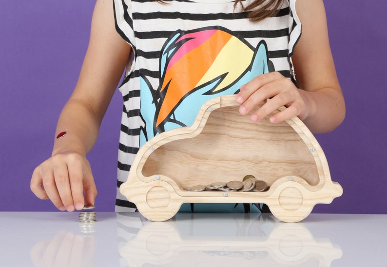 May include: A wooden car-shaped money box with a slot on top and a clear front. The box is filled with coins. The car is made of light-coloured wood and has a simple design. The background is a solid purple.