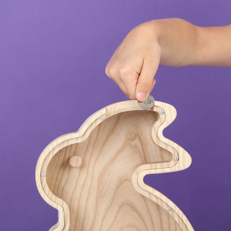May include: A light-coloured wooden bunny-shaped money box with a coin slot. The money box is made of light-coloured wood and has a smooth finish. A hand is placing a coin into the slot. The background is a solid purple colour.