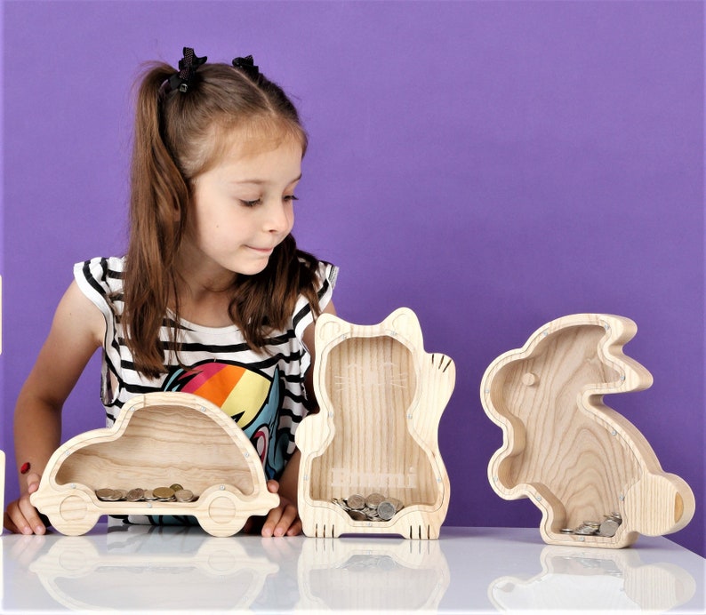 May include: Three wooden coin banks shaped like a car, cat, and rabbit, each filled with coins. They are on a white surface against a purple backdrop. The car bank is rectangular with wheels. The cat bank has a cat face. The rabbit bank is shaped like a bunny.