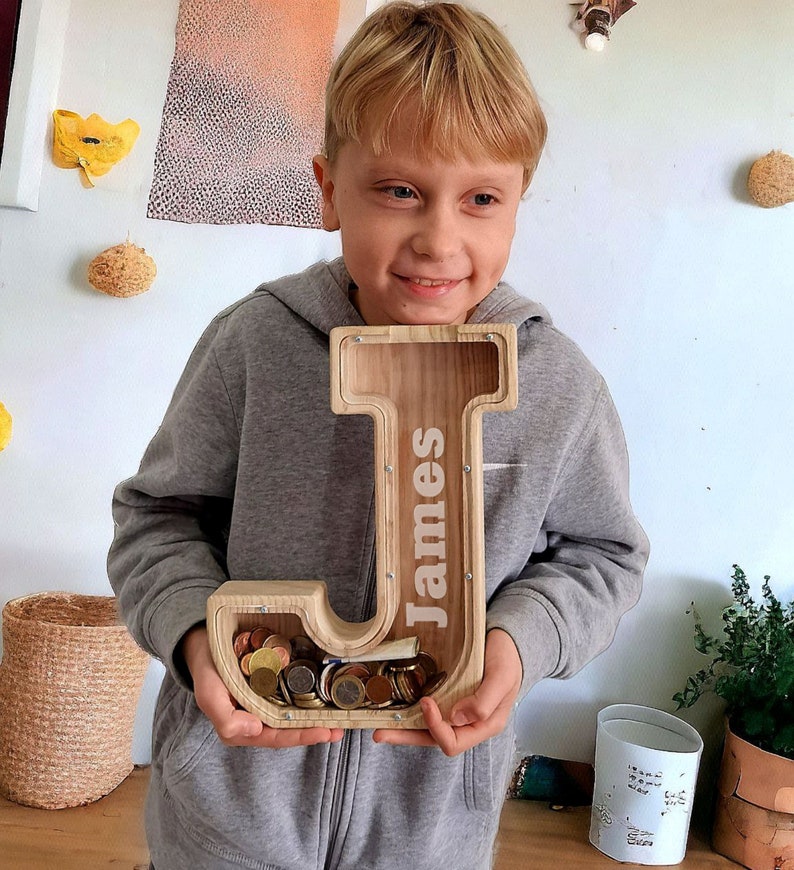 Op de afbeelding: Een houten spaarpot in de vorm van de letter J met de naam "James" erop gegraveerd. De spaarpot zit vol met munten en wordt vastgehouden door een kind.