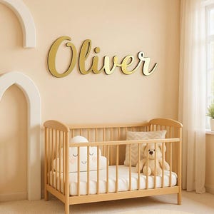 Puede incluir: Una escena de guardería con una cuna de madera, una almohada en forma de nube y un oso de peluche. El nombre "Oliver" se muestra en letras doradas en la pared sobre la cuna. La habitación tiene una paleta de colores neutros.