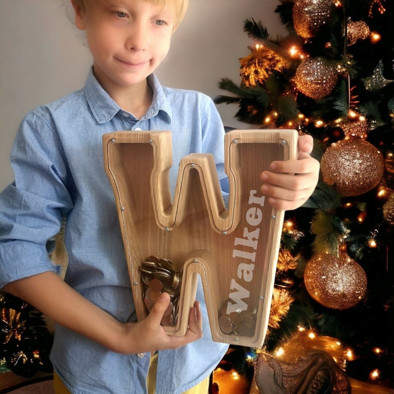K&ouml;nnte beinhalten: Ein Sparschwein aus Holz in Form des Buchstabens W mit einem Schlitz f&uuml;r M&uuml;nzen. Der Buchstabe ist mit dem Namen "Walker" personalisiert.