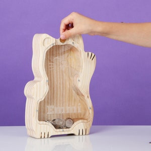 May include: A wooden cat-shaped money box with a clear front, containing coins. The bank has a slot at the top for inserting coins and the name "Danni" is visible. A hand is placing a coin into the slot.