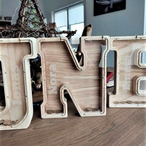 K&ouml;nnte beinhalten: Holzbuchstaben-Sparschwein mit den Buchstaben "ONE" ausgeschrieben. Die Buchstaben sind in einer hellen Holzfarbe lackiert und haben oben einen Schlitz f&uuml;r M&uuml;nzen. Der Name "Nora" ist auf den Buchstaben "N" geschrieben.