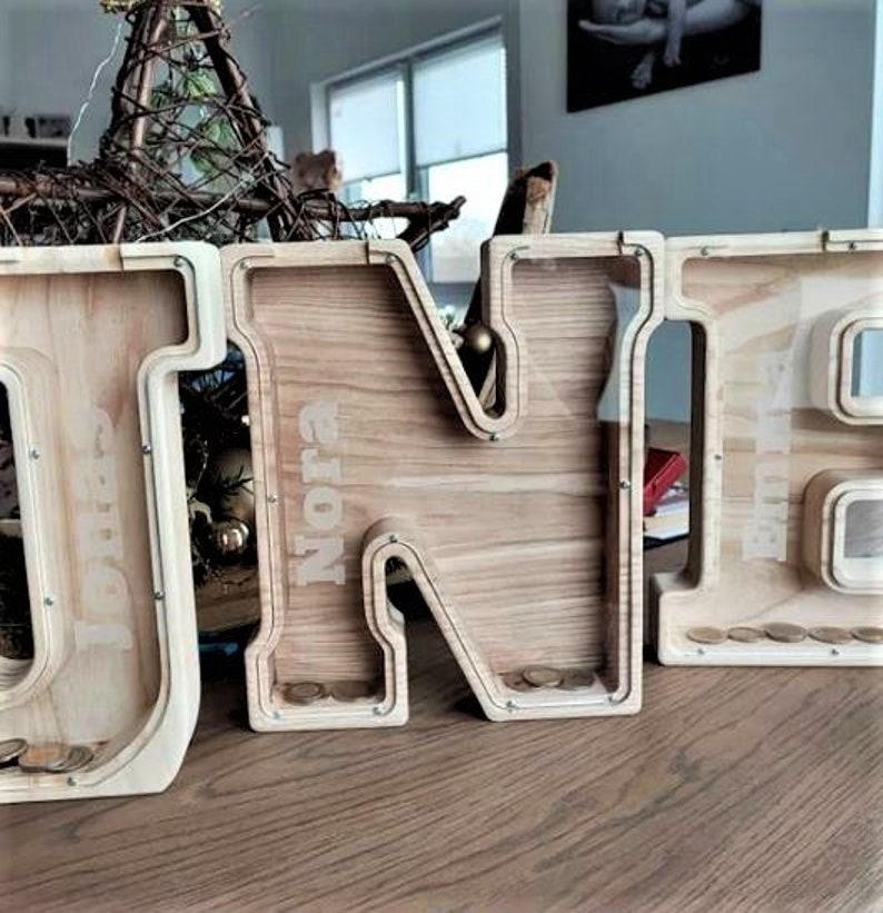 K&ouml;nnte beinhalten: Holzbuchstaben-Sparschwein mit den Buchstaben "ONE" ausgeschrieben. Die Buchstaben sind in einer hellen Holzfarbe lackiert und haben oben einen Schlitz f&uuml;r M&uuml;nzen. Der Name "Nora" ist auf den Buchstaben "N" geschrieben.