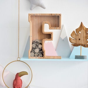 K&ouml;nnte beinhalten: Holzbuchstaben F Sparschwein mit einer durchsichtigen Frontplatte, gef&uuml;llt mit M&uuml;nzen. Das Sparschwein steht auf einem hellblauen Regal mit einer rosa und blauen Bergdekoration, einer Holzblattdekoration und einer goldenen Kette, die von der Decke h&auml;ngt.