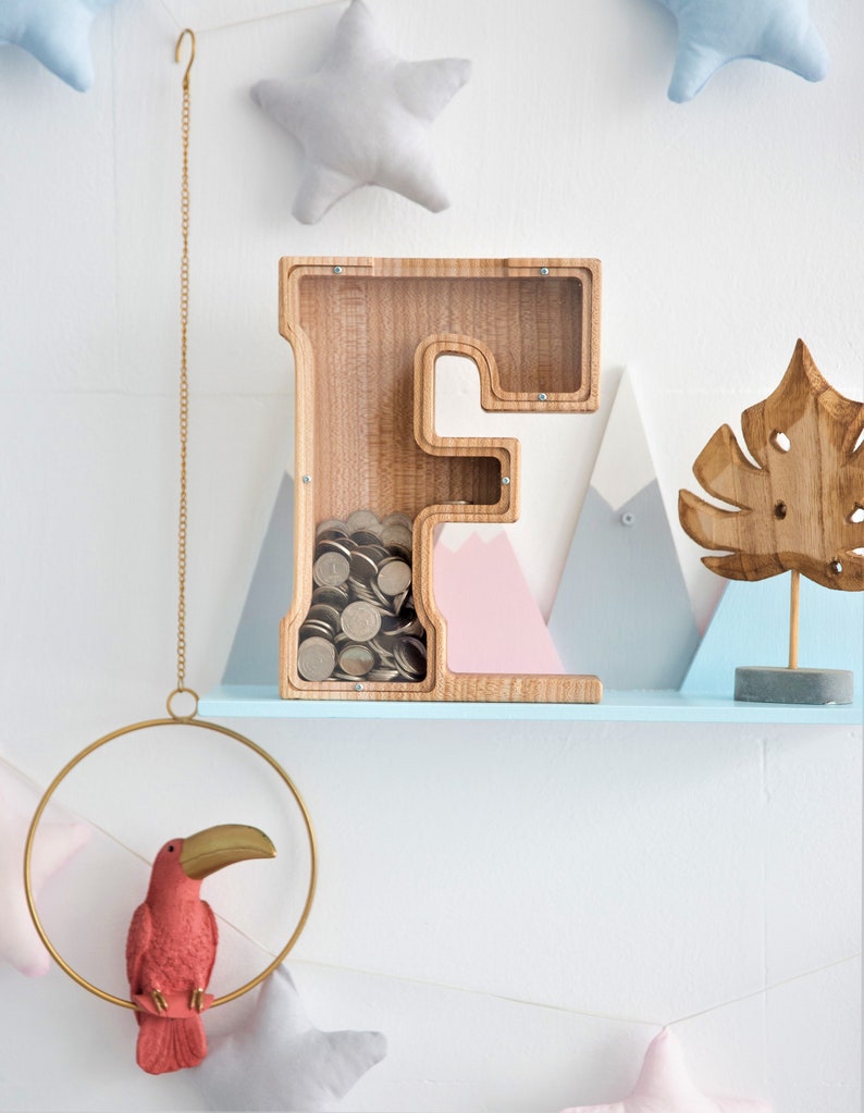 K&ouml;nnte beinhalten: Holzbuchstaben F Sparschwein mit einer durchsichtigen Frontplatte, gef&uuml;llt mit M&uuml;nzen. Das Sparschwein steht auf einem hellblauen Regal mit einer rosa und blauen Bergdekoration, einer Holzblattdekoration und einer goldenen Kette, die von der Decke h&auml;ngt.