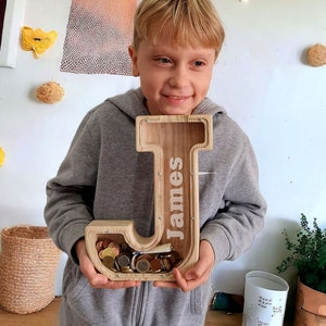 May include: A wooden piggy bank shaped like the letter J with the name "James" engraved on it. The piggy bank is filled with coins and is being held by a child.