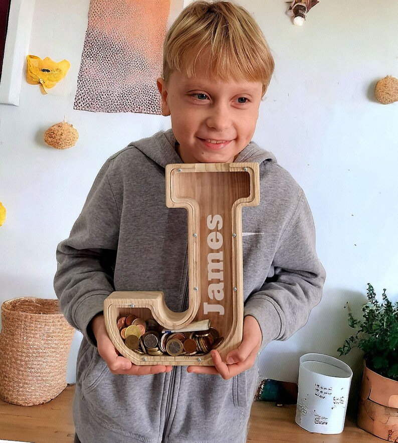 May include: A wooden piggy bank shaped like the letter J with the name "James" engraved on it. The piggy bank is filled with coins and is being held by a child.