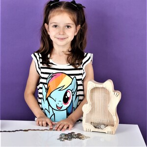 May include: A wooden cat-shaped money box with a clear front, containing coins. The money box is on a white surface. A young person wearing a black and white striped top with a cartoon character graphic is standing next to the money box.