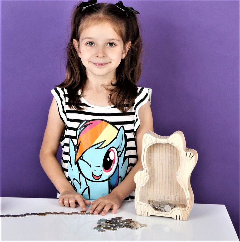 May include: A wooden cat-shaped money box with a clear front, containing coins. The money box is on a white surface. A young person wearing a black and white striped top with a cartoon character graphic is standing next to the money box.