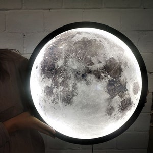 May include: A black round moon lamp with a white moon image. The moon lamp has a white light that illuminates the moon image.