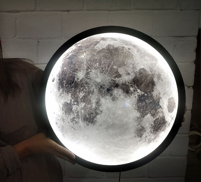 May include: A black round moon lamp with a white moon image. The moon lamp has a white light that illuminates the moon image.