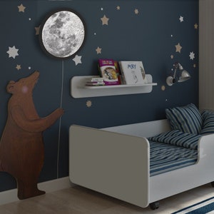 May include: A white toddler bed with blue and white striped bedding. The bed has a white headboard and footboard and is on wheels. The wall behind the bed is blue and has white stars and a brown bear sticker. A white shelf with books is mounted on the wall. A moon-shaped nightlight is on the wall above the bear sticker.