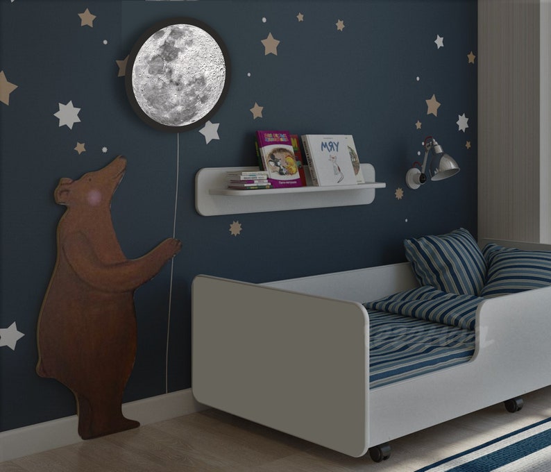 May include: A white toddler bed with blue and white striped bedding. The bed has a white headboard and footboard and is on wheels. The wall behind the bed is blue and has white stars and a brown bear sticker. A white shelf with books is mounted on the wall. A moon-shaped nightlight is on the wall above the bear sticker.