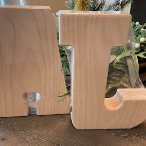 K&ouml;nnte beinhalten: Zwei unfertige Holzbuchstaben, A und J, liegen auf einem Holztisch. Die Buchstaben sind aus hellem Holz gefertigt und haben eine glatte, geschliffene Oberfl&auml;che.