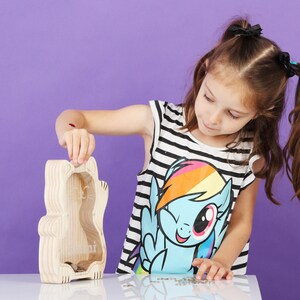 May include: A wooden cat-shaped money box with a clear front, shown with coins inside. A person is putting a coin into the slot. The person is wearing a black and white striped top with a colourful graphic.