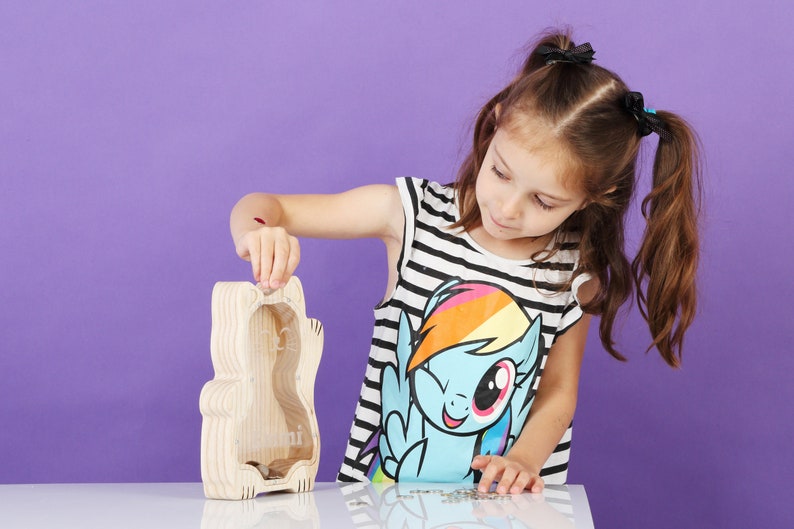 May include: A wooden cat-shaped money box with a clear front, shown with coins inside. A person is putting a coin into the slot. The person is wearing a black and white striped top with a colourful graphic.