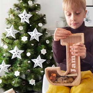 May include: A wooden letter "J" shaped piggy bank with the name "James" printed on it. The bank is filled with coins. A Christmas tree with white star ornaments is in the background.