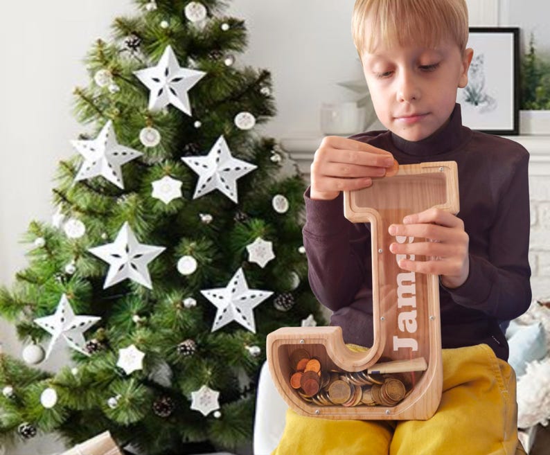 May include: A wooden letter "J" shaped piggy bank with the name "James" printed on it. The bank is filled with coins. A Christmas tree with white star ornaments is in the background.