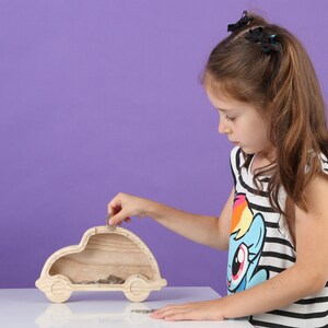 May include: A wooden car-shaped money box with a slot on top. A child is putting a coin into the slot. The money box is made of light-coloured wood and sits on a white surface against a purple background. The child wears a black and white striped top.