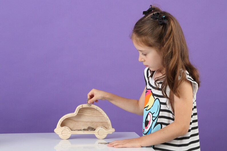 May include: A wooden car-shaped money box with a slot on top. A child is putting a coin into the slot. The money box is made of light-coloured wood and sits on a white surface against a purple background. The child wears a black and white striped top.