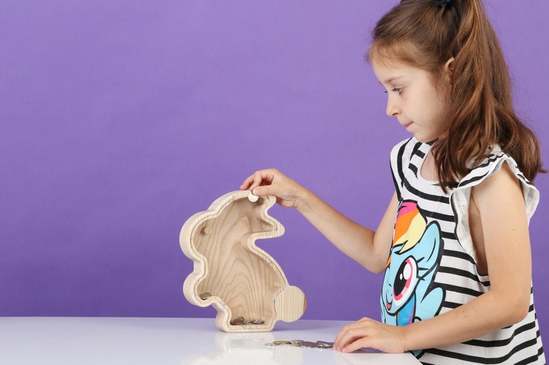 May include: A wooden bunny-shaped money box with a slot on top and a coin slot on the back. The box is made of light-coloured wood and has a simple design. The money box is on a white surface.