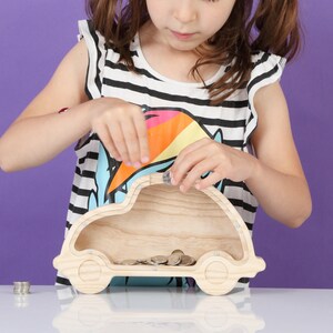 May include: A wooden car-shaped money box with coins inside. The bank is light-coloured and has a slot on top for inserting coins. The image shows a child adding coins to the bank. The background is a solid purple.