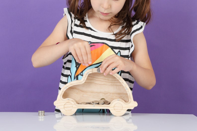 May include: A wooden car-shaped money box with coins inside. The bank is light-coloured and has a slot on top for inserting coins. The image shows a child adding coins to the bank. The background is a solid purple.