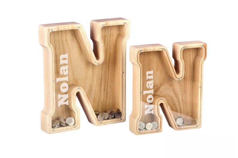 May include: Two wooden money boxes shaped like the letter N. Each box has the name Nolan engraved on the front. The boxes are filled with coins.