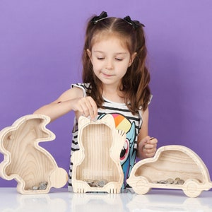 May include: Three wooden coin banks shaped like a rabbit, a bear, and a car. The banks are made of light-coloured wood and have slots for coins. A young person is shown interacting with the banks.