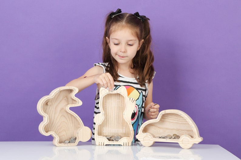May include: Three wooden coin banks shaped like a rabbit, a bear, and a car. The banks are made of light-coloured wood and have slots for coins. A young person is shown interacting with the banks.