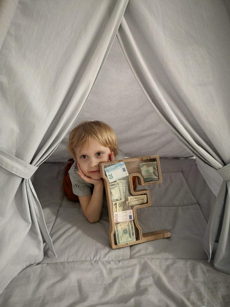 May include: A wooden letter "E" shaped money bank filled with various paper currency. The bank is open, revealing the contents. A young child is resting their head on their hand, looking at the bank. The scene is set inside a gray tent.