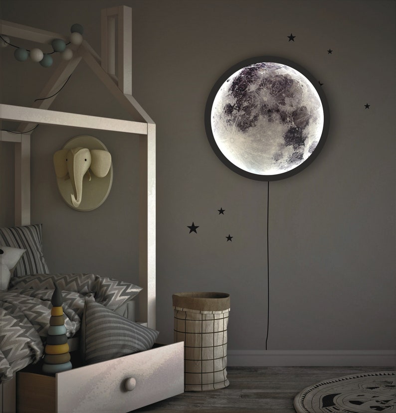 May include: A white wooden bed frame with a gray and white patterned comforter and pillows. A wooden toy stacking tower sits on the bed. A round moon-shaped nightlight with a black frame hangs on the wall. A woven basket sits on the floor next to the bed.