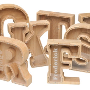 May include: Wooden letter piggy banks with names "Ridge" and "Frederick" spelled out. The letters are made of light brown wood and have a clear plastic front.